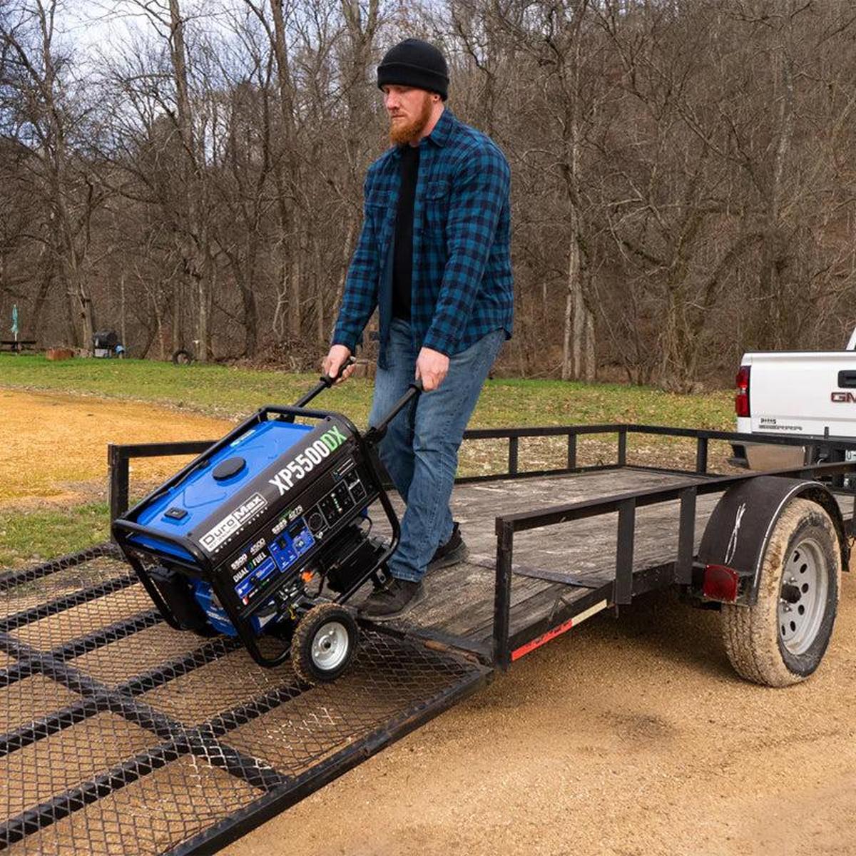 A man in a blue plaid shirt and black beanie is loading a blue and black Duromax XP5500DX portable generator onto a trailer in a grassy outdoor area, demonstrating the generator's ease of transport.